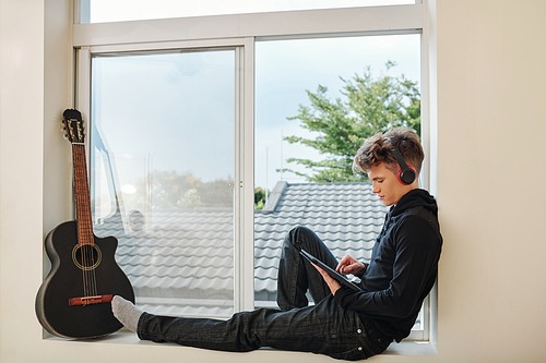 Teenage boy sitting on window sill with headphones and watching webinar or movie on tablet computer