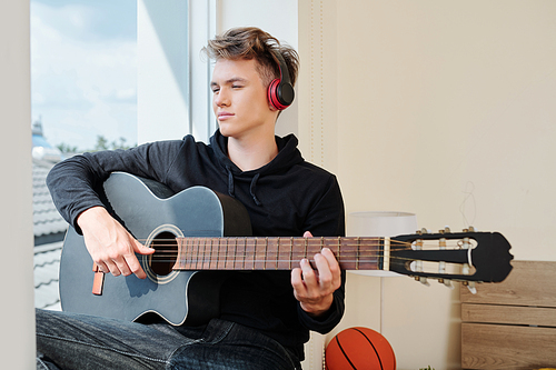 Portrait of pensive teenager in headphones working on new song when sitting on window sill of his room