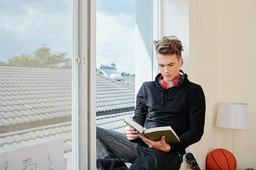 Serious teenage boy in jeans and hoodie sitting on window sill and reading interesting book