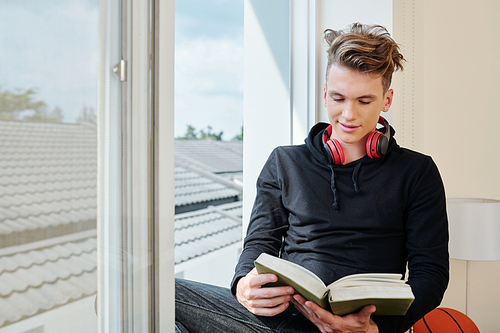 Positive teenage boy sitting at window of his room and reading new captivating book