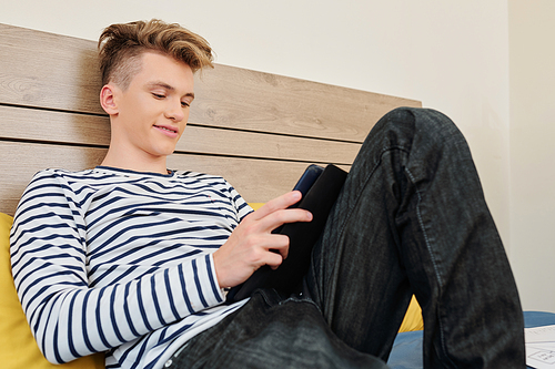 Smiling teenage boy resting in his bedroom and watching sitcom on tablet computer