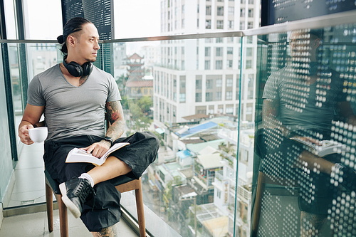 Pensive young man with openend book drinking coffee and looking at big city from the balcony of his apartment