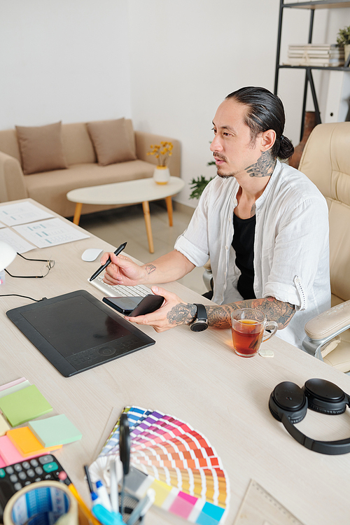 Experienced designer with smartphone in hands working on graphic tablet at his home office