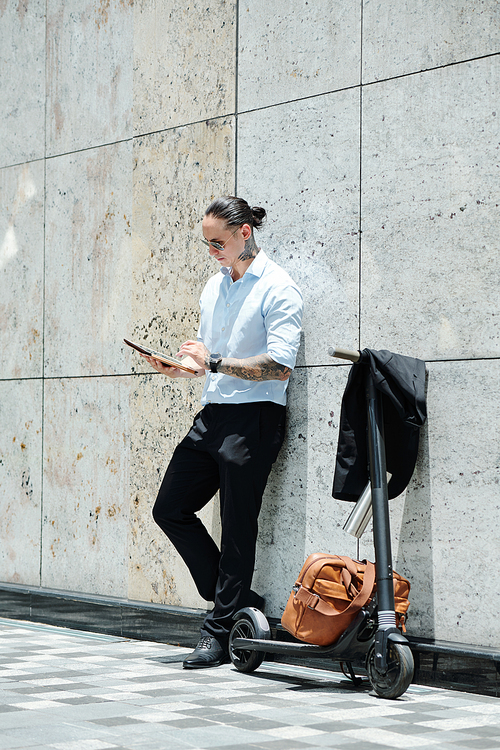 Young fashionable businessman standing outdoors next to his scooter and reading document on digital tablet