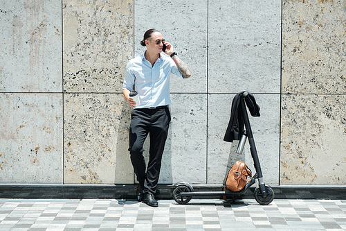 Stylish confident young businessman leaning on wall, drinking take out and talking on phone