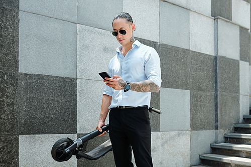 Serious frowning young businessman in glasses holding scooter and answering text messages from colleagues and clients