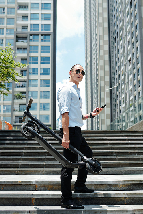 Young man in sunglasses standing on steps with scooter and smartphone in hands and looking away