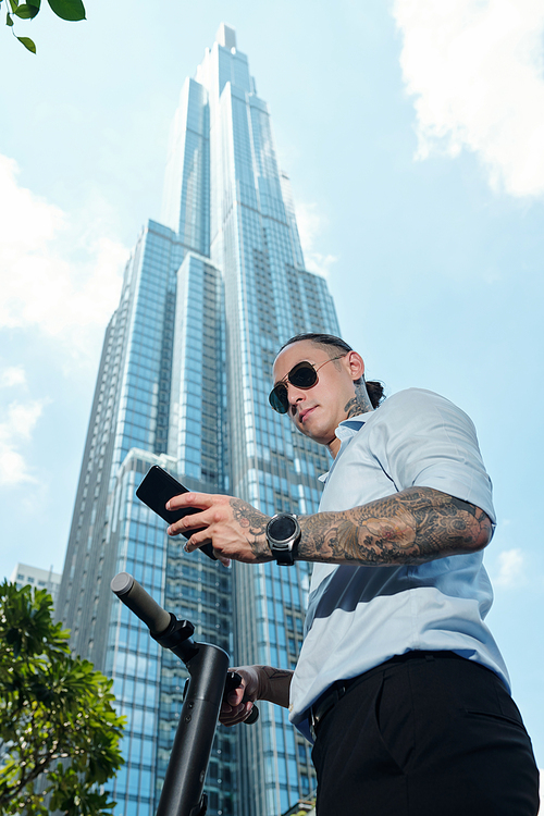 Serious young businessman standing on scooter in front of sckyscraper and reading text message on smartphone