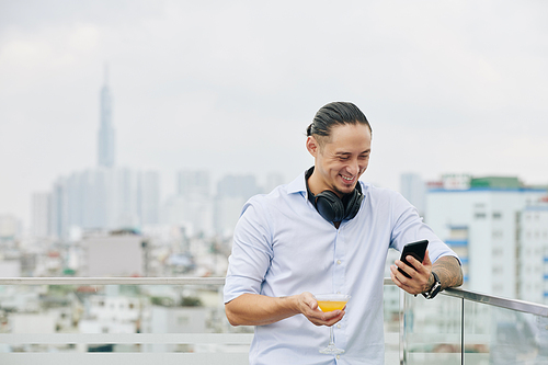 Laughing handsome young man standing on rooftop with glass of fruit cocktail and watching funny video on smartphone