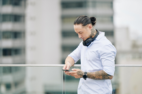 Portrait of pensive mixed-race man standing on balcony and checking text messages on smartphone