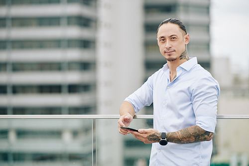 Portrait of handsome young mixed-race man with smartphone standing on rooftop and looking at camera