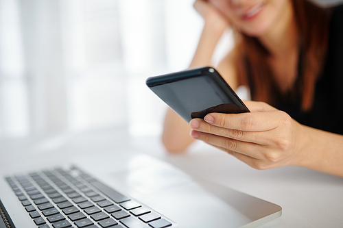 Close-up image of smiling young woman sitting at office desk and checking social media during short break