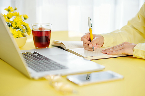 CLose-up image of female student watching webinar on laptop at home and taking notes in notebook