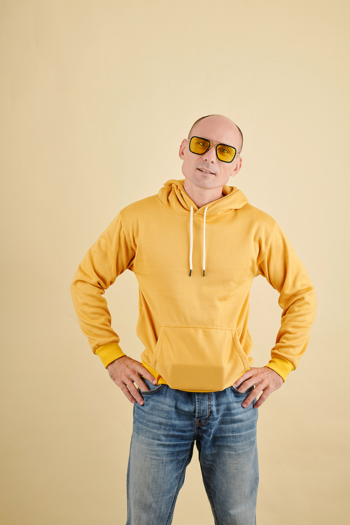 Studio portrait of confident middle-aged man in bright yellow hoodie standing with hand on hips and looking at camera