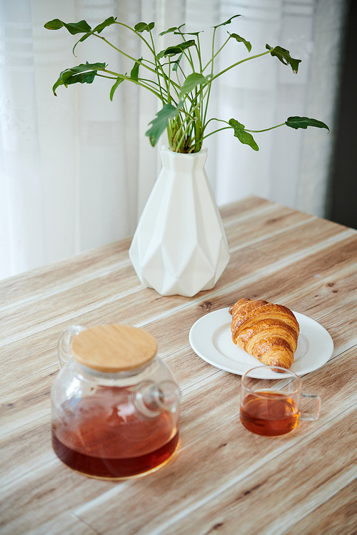 Teapot, cup and fresh croissant on kitchen table served for breakfast