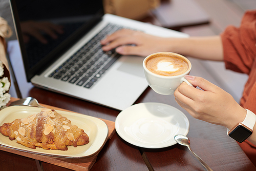 Hands of woman having cup of cappuccino and fresh croissant for breakfast and checking e-mails on laptop