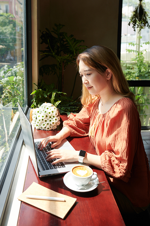 Smiling young woman sitting at coffeeshop windown and rays of sun and working on laptop