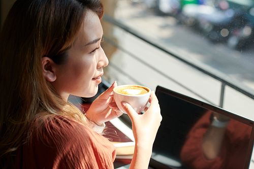 Smiling woman enjoying morning coffee in coffeeshop and looking on through big window