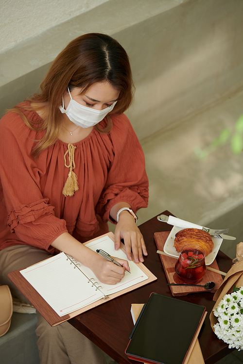 Young woman in medical mask eating breakfast in cafe and writing down plans and ideas in notepad
