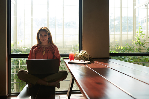 Cheeful young woman reading artciles on laptop screen when sitting at big cafe table