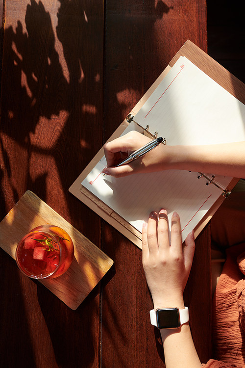 Hands of creative young woman drinking fruit cocktail and writing in planner, view from the top