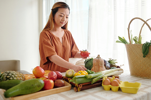 Smiling pretty young woman putting fresh ripe vegetables from her garden on trays on kitchen table