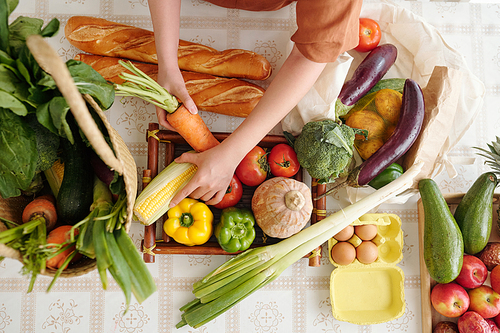 Hands of young woman taking out fresh bread, fruits and vegetables she bought in supermarket on kitchen table