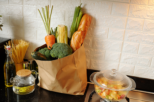 Paper bag of fresh groceries on kitchen counter next to teapot, bottle of olive oil and stove with boiling pasta