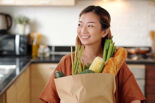 Portrait of happy pretty young woman standing in kitchen with paper bag of fresh groceries like bread and vegetables