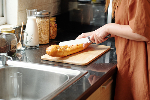 Woman standing at kitchen counter and cutting fresh baguette with sharp knife