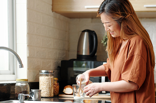 Pretty smiling young woman pouring fresh milk in glass on kitchen counter