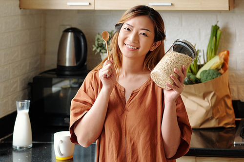 Portrait of happy young woman with jar of oatmeal and wooden spoon standing in kitchen