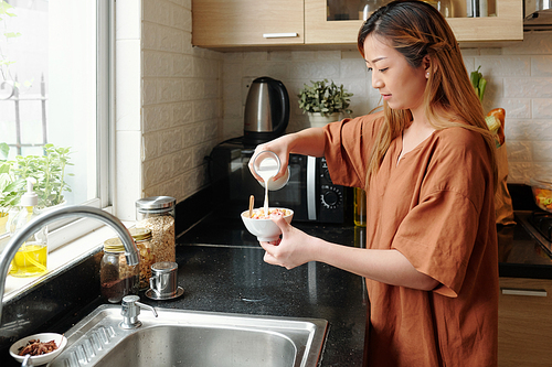 Serious pretty young woman pouring milk in bowl with granola and berries