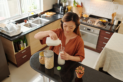 Pretty young woman standing at kitchen counter and pouring milk in glass jar