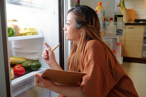 Pensive woman with notepad checking groceries in her fridge and writing down a list of products she need to buy