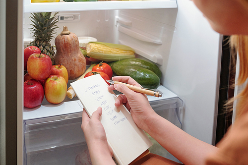 Hands of woman writing down list of groceries she needs to buy in supermarket or order online