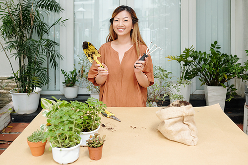 Portrait of happy young smiling woman standing at big table with trowel and gardening fork in hands ready for reporting flowers