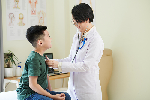 Smiling pediatrician with stethoscope listening to heartbeat of cheerful preteen boy