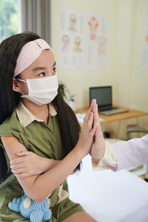 Little girl in medical mask giving high five to general practitioner after getting vaccine in hospital