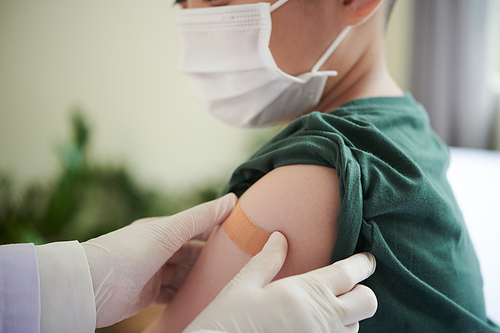 Doctor sticking adhesive plaster over covid-19 vaccine injection site on arm of preteen boy in medical mask