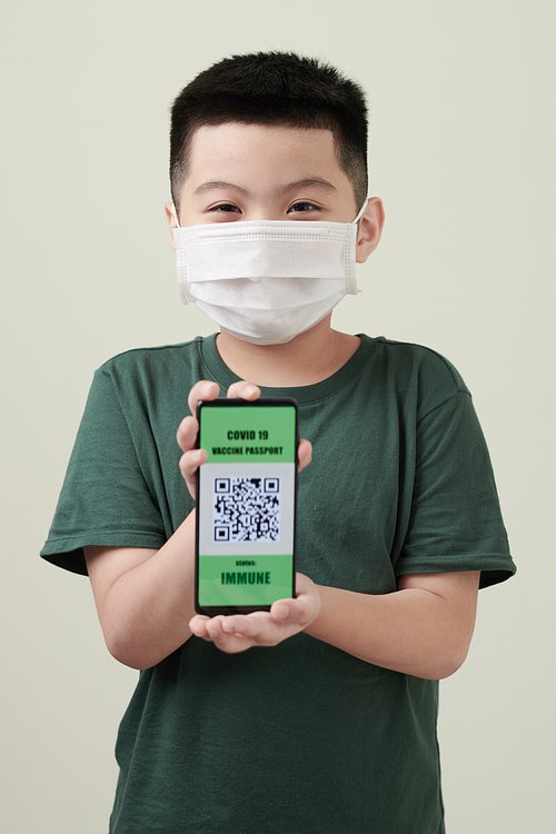 Portrait of happy little boy in medical mask showing smartphone with immune status on screen