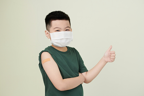 Cheerful little boy wearing medical mask and showing thumbs-up after getting shot of vaccine in arm