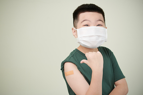 Emotional excited little boy in medical mask showing adhesive plaster over his covid-19 vaccine injection site
