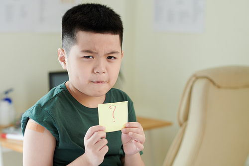 Frowning doubting little boy with adhesive plaster on shoulder showing sticker note with question mask