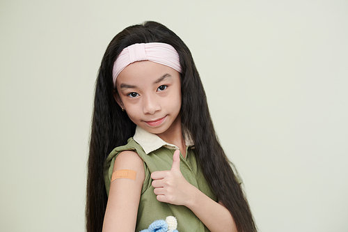 Portrait of positive preteen girl with adhesive plaster over her coronavirus vaccine injection site showing thumbs-up