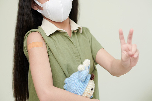 Little girl in protective mask having adhesive plaster on her shoulder and showing peace gesture