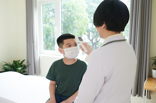 Curious little boy in medical mask looking at pediatrician measuring body temperature with non-contact infrared thermometer