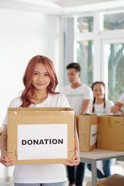 Young Asian woman with pink hair holding big box of groceries and clothes people donated at charitable foundation