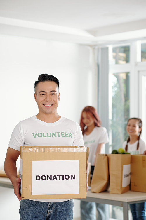Smiling handsome young Vietnamese volunteer holding big cardboard box of donated groceries
