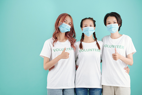 Young female volunteers in protective masks hugging and showing thumbs-up